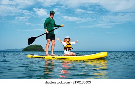 A Father Rides His Little Daughter In A Life Jacket On A Big Yellow SUP Board In The Sea. Active Outdoor Recreation For The Whole Family.