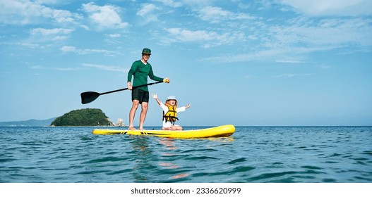 A father rides his little daughter in a life jacket on a big yellow SUP board in the sea. Active outdoor recreation for the whole family.