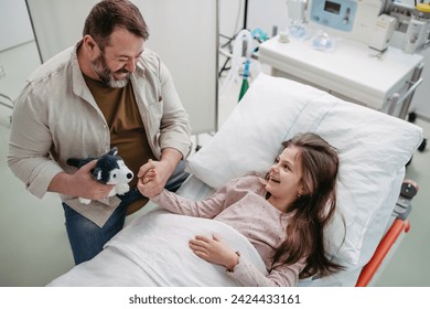 Father sitting beside daughter lying on hospital bed, playing with plush toy. Gift from dad to sick little girl in hospital. - Powered by Shutterstock