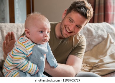 Father Sitting On The Edge Of The Sofa In The Living Room And Burping Her Newborn Child After Feeding Him. 