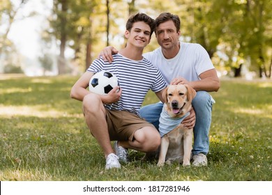 father sitting on grass near pleased teenager son with football and golden retriever