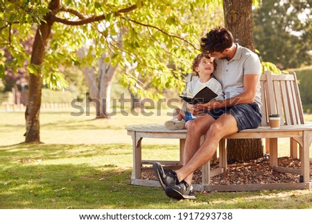 Father Sitting On Park Bench Under Tree With Son Reading Book Together