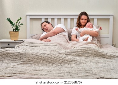 Father Sleeps While Mother Takes Care Of Baby Boy, Parents And Infant Child On Home Bed. Problems Of A Man And A Woman With A Newborn Baby In The Bedroom