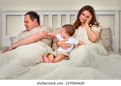 Father Sleeps While Mother Takes Care Of Baby Boy, Parents And Infant Child On Home Bed. Problems Of A Man And A Woman With A Toddler Baby In The Bedroom