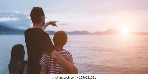 Father With Son And Daughter Near River At Sunset. Dad And Kids Watching Sunrise Together. Happy Man And Children Relaxing On Beach, Family Enjoying Beautiful View. Bonding And Togetherness