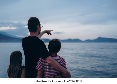 Father With Son And Daughter Near River At Sunset. Dad And Kids Watching Sunrise Together. Happy Man And Children Relaxing On Beach, Family Enjoying Beautiful View. Bonding And Togetherness