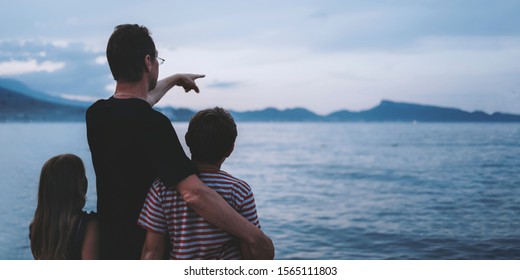 Father With Son And Daughter Near River At Sunset. Dad And Kids Watching Sunrise Together. Happy Man And Children Relaxing On Beach, Family Enjoying Beautiful View. Bonding And Togetherness