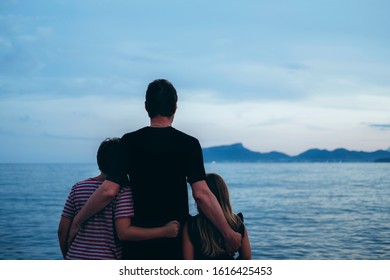 Father With Son And Daughter Near River At Sunset. Dad And Kids Watching Sunrise Together. Happy Man And Children Relaxing On Beach, Family Enjoying Beautiful View. Bonding And Togetherness