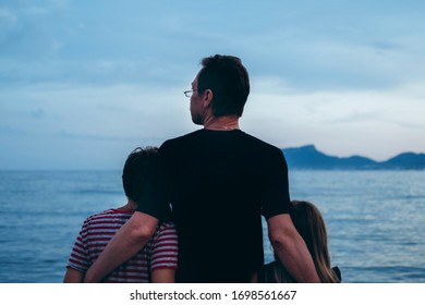 Father With Son And Daughter Near River At Sunset. Dad And Kids Watching Sunrise Together. Happy Man And Children Relaxing On Beach, Family Enjoying Beautiful View. Bonding And Togetherness