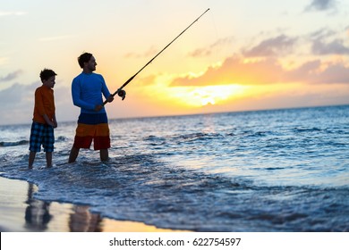 Father And Son Fishing Together In Ocean From Beach On Sunset