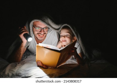 Father and son with flashlight reading book under blanket at home - Powered by Shutterstock