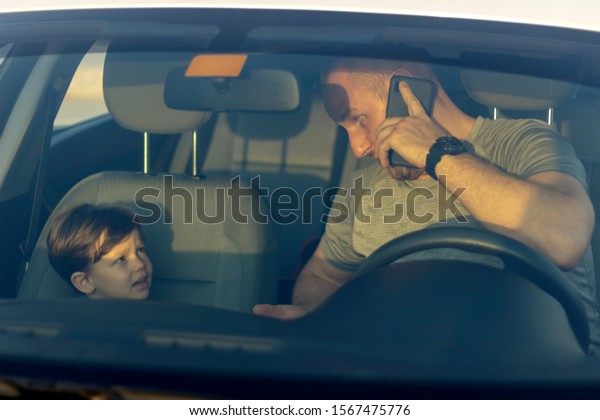 Father and son going on the road trip and having fun\
in the car.Adorable little boy smiling at camera while father\
talking on smartphone in car.Dad using mobile while his little boy\
looking at him.