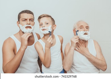 Father, son and grandfather standing with shaving cream on their faces shaving with razors, isolated on white
