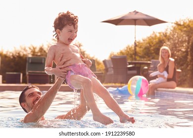 Father And Son Have Fun Playing In Outdoor Pool On Vacation As Mother And Baby Watch From Side