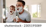 Father and son having fun in a bathroom, laughing happily with shaving foam on their faces. Young single dad taking a moment to bond and share moments of joy with his boy on father