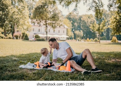 Father And Son Having A Play Date In The Park, Sitting On A Blanket In The Grass. They Are Celebrating International Father’s Day.