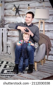 Father And Son. A Man Sitting In A Chair And Next To Him A Small Child 2-3 Years Old Against A Wooden Wall. A Young Dark-haired Man With A Child In A Christmas Decoration.