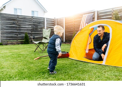 Father and son play camp in the backyard. Young father and his little toddler boy are playing together in an orange tent outdoors. Folding chair, guitar are near on the lawn