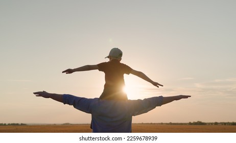 Father Son play together in front of sun, dream, fly. Dad, child fantasize, kid aviator sits on his fathers shoulders. Concept of happy family, childhood dream. Boy plays pilot airplane, hands wings - Powered by Shutterstock