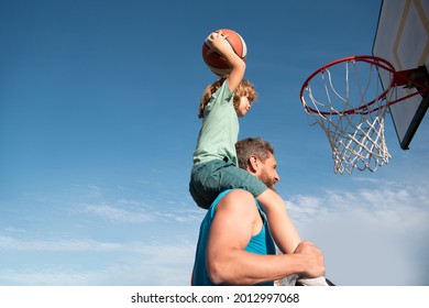 Father And Son Playing Basketball. Sports Concept. Boy Child Sitting On The Dad Shoulders, Throwing Basketball Ball Into Basket, Side View On Sky Background With Copy Space.