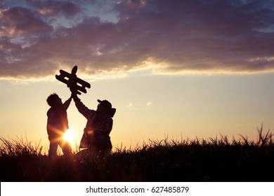 Father And Son Playing With Cardboard Toy Airplane In The Park At The Sunset Time. Concept Of Friendly Family. People Having Fun Outdoors. Picture Made On The Background Of Dark Blue Sky.