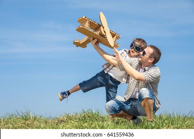 Father And Son Playing With Cardboard Toy Airplane In The Park At The Day Time. Concept Of Friendly Family. People Having Fun Outdoors. Picture Made On The Background Of Blue Sky.