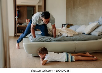 Father and son playing hide and seek in bedroom. Little boy hiding by the bed with father searching him. Family playing games inside their home.