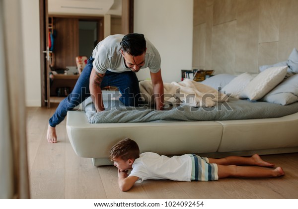 Father and son playing hide and seek in bedroom.\
Little boy hiding by the bed with father searching him. Family\
playing games inside their\
home.