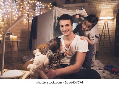 Father and son are playing with little baby sister in blanket fort at night at home.