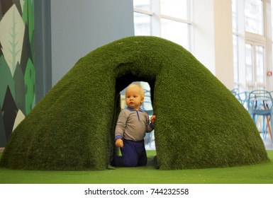 Father And Son Are Playing On Artificial Grass. The Concept Of A Friendly Family. A Blond Child Hides In An Artificially Created Tunnel From Grass. Dad Runs After His Son. Special Vintage Processing