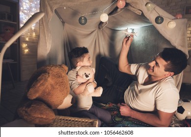 Father and son are playing with toy airplane and cars in blanket fort at night at home.