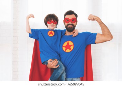 Father and son in the red and blue suits of superheroes. On their faces are masks and they are in raincoats. They are posing in a bright room.
