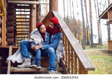 father and son in red hats and knitted scarves are sitting on the porch stairs at a wooden house made of logs in autumn - Shutterstock ID 2060412914