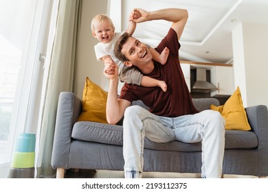 Father And Son Relationships. Handsome Dad Carrying Baby Boy On His Shoulders, Bending To Side Pretending To Fall Making Cute Kid Laugh With Excitement Sitting On Grey Sofa. Fatherhood, Parenting