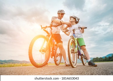 Father And Son Ride, Cycling Their Bikes Together, On Sunny Day, Happy Family In Helmets Is Riding Bikes Having Fun.