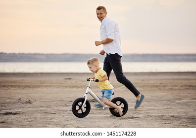 Father with son riding bicycle in summer. Man runs along with kid on the beach at sunset. Son learns to ride a bike