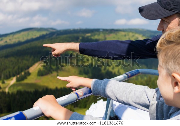 Father and son riding up to\
the mountain in chairlift and pointing at something in the\
distance. 