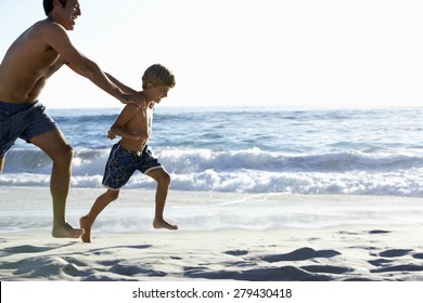 Father And Son Running Along Beach Together Wearing Swimming Costumes