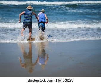 Father And Son Are Running Towards Waves. Blue Clothes Are Matching Blue Sea In Background.