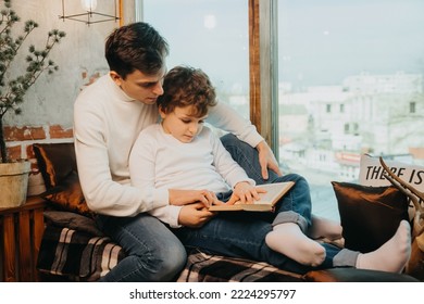 Father and son in the same white sweaters are sitting on a window sill and reading a fairy tale book.
 - Powered by Shutterstock