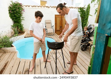 Father and son setting up a barbecue grill in a cozy backyard by the pool. Family time, outdoor activity, and bonding on a sunny day. - Powered by Shutterstock