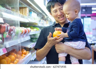 Father And Son Are Shopping In A Supermarket. Smiling Dad And Cute Little Asian 12 Months / 1 Year Old Baby Boy Child Choosing Fruits In Grocery Store, Kid First Experience Concept