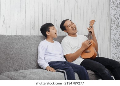 Father and son are singing together while playing ukulele, wearing white t shirt sitting on sofa, playing some music at home  - Powered by Shutterstock