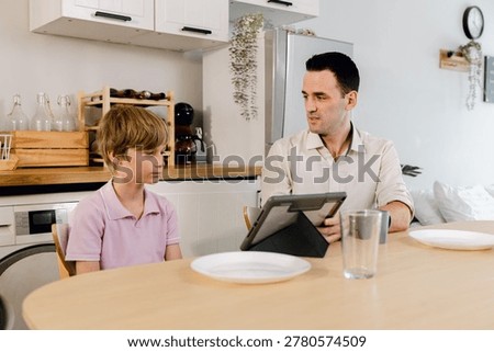 A father and son sit at a kitchen table, engaged in a serious conversation with a tablet in front of them.