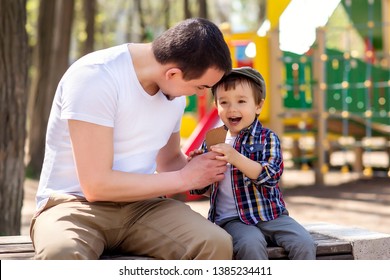 Father And Son Sit On Bench And Eat Ice Cream In Park In Sunny Spring Or Summer Day. Both Dad And Child Are Smiling, Playground In Background. Family Fun Outdoor And Father And Son Spend Time Together