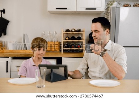 A father and son are sitting at a kitchen table, engaging with a tablet while the father enjoys a cup of coffee.