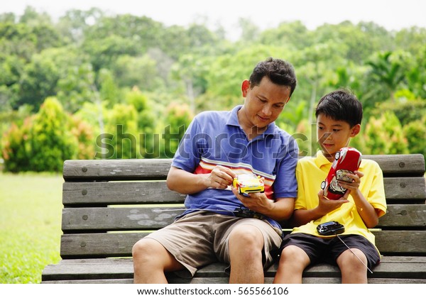 Father and son, sitting on bench, holding remote\
control cars