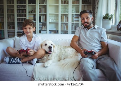 Père et fils assis sur un canapé avec un chien de compagnie et jouant à des jeux vidéo à la maison : photo de stock