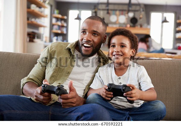 Father And Son Sitting On Sofa In Lounge Playing\
Video Game