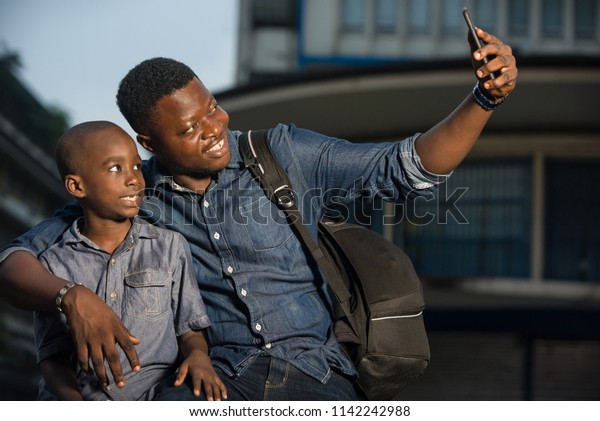 Father and son sitting in the street waiting\
for a car, take a photo with a smart phone. concept of tourist and\
travel with children.